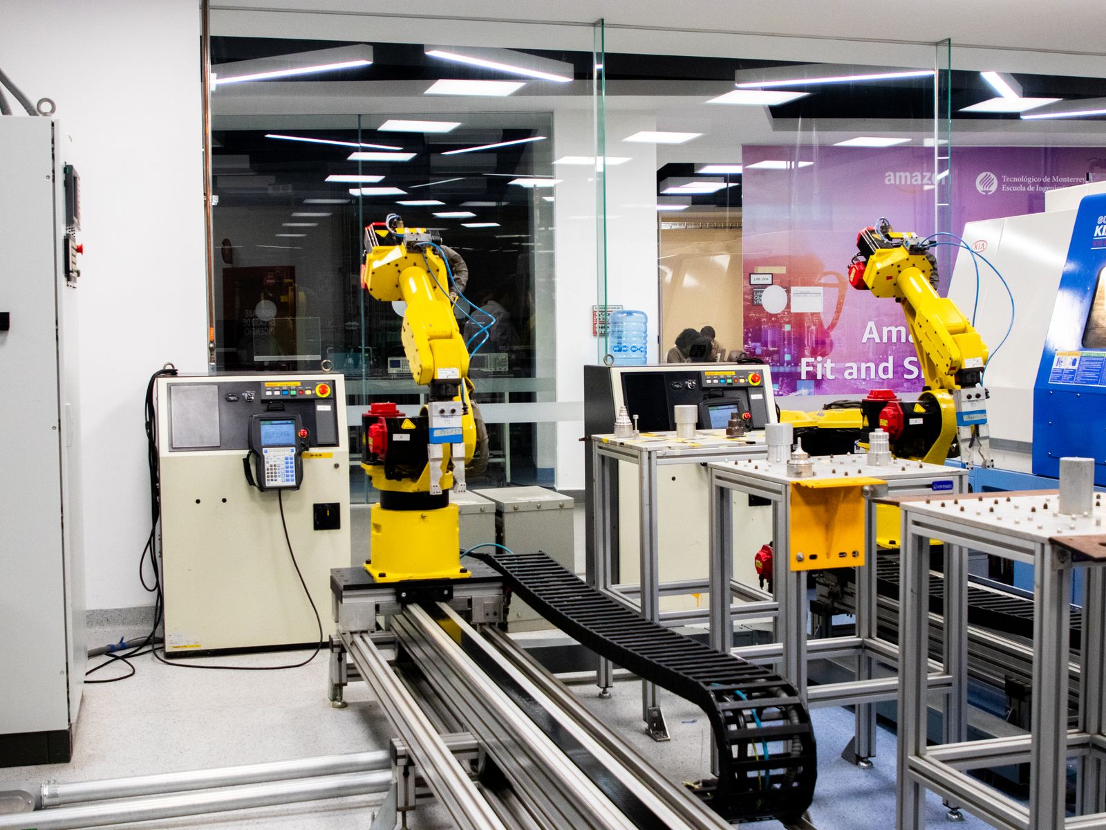 Two yellow robot arms beside control cabinets, teach pendants and component fixtures.